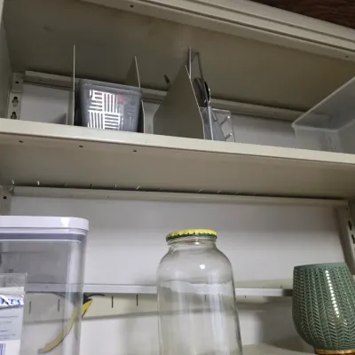 Sale Photo Thumbnail #135: Assorted household storage items, including a glass jar, plastic containers, and a green textured vase, arranged on metal shelving.