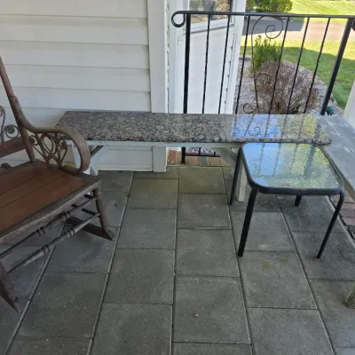 Sale Photo Thumbnail #212: Outdoor patio furniture set featuring an antique wooden rocking chair with metal scrollwork, a long bench with a polished granite top, a rustic wooden bench, and a small glass-top side table.