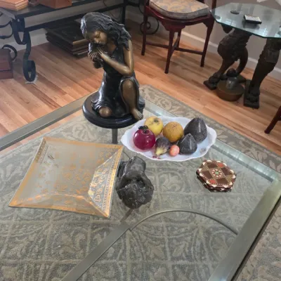 Sale Photo Thumbnail #50: Assorted tabletop decor including a bronze-finish kneeling girl statue, a gold-patterned glass tray, a bowl of decorative stone fruit, and a set of inlaid wooden coasters on a glass-top coffee table.