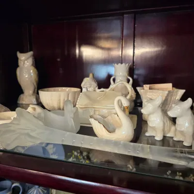 Sale Photo Thumbnail #34: Collection of vintage white and iridescent ceramic figurines, including an owl, swans, and dogs, displayed alongside frosted glass items on a cabinet shelf.