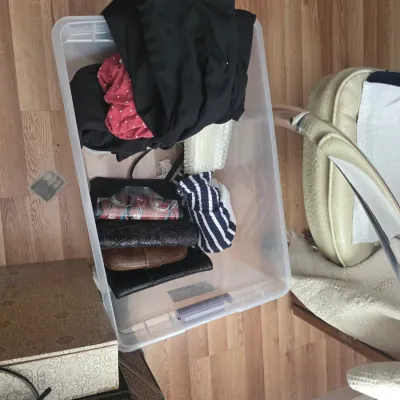 Sale Photo Thumbnail #114: Clear plastic storage bin containing an assortment of clothing items and small handbags, shown on a wood-look floor.