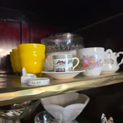 Sale Photo Thumbnail #22: Assorted vintage glassware and teacups, including a bright yellow tumbler, a small patterned demitasse cup with saucer, and floral porcelain cups displayed on a shelf.