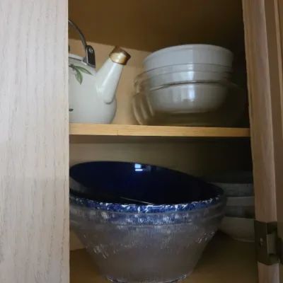 Sale Photo Thumbnail #130: Assorted kitchenware stored in a wooden cabinet, including a white ceramic teapot, glass mixing bowls, and a cobalt blue bowl.
