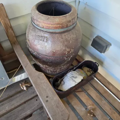 Sale Photo Thumbnail #217: Large, rustic glazed stoneware crock with a textured brown finish, shown alongside a small metal trough on a wooden bench.