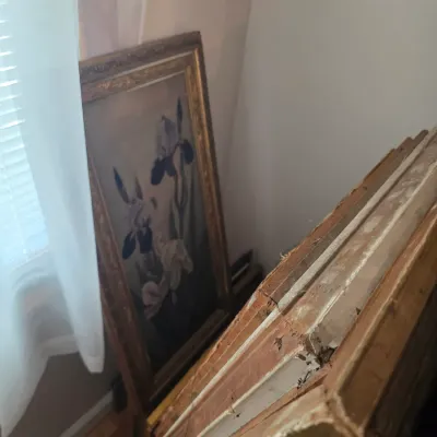Sale Photo Thumbnail #45: Vintage framed oil painting of irises in a gilded wood frame, shown leaning against a wall next to a stack of other framed artworks.