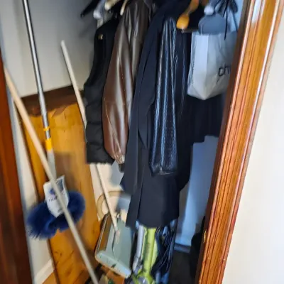 Sale Photo Thumbnail #157: Assorted outerwear hanging in a closet, including a brown leather jacket and black coats, alongside cleaning supplies like a broom, mop, and vacuum cleaner.