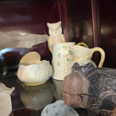 Sale Photo Thumbnail #33: Collection of vintage Belleek porcelain pieces, including a shamrock-patterned pitcher, an owl figurine, and a shell-shaped dish, displayed on a glass shelf.