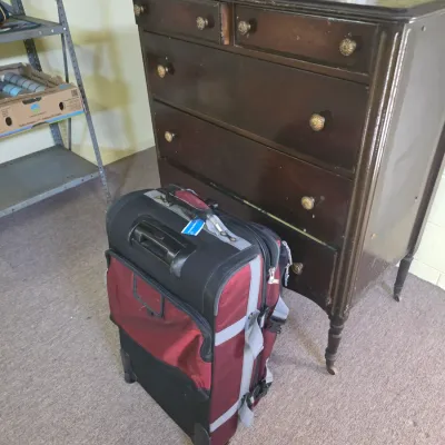 Sale Photo Thumbnail #190: Vintage dark wood five-drawer dresser with brass knobs and turned legs on casters, shown next to a maroon and black rolling suitcase.
