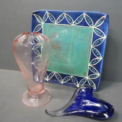 Sale Photo Thumbnail #17: Art glass and ceramic collection featuring a pink glass vase with a scalloped rim, a cobalt blue freeform glass sculpture, and a square ceramic platter with a teal center and patterned blue border.