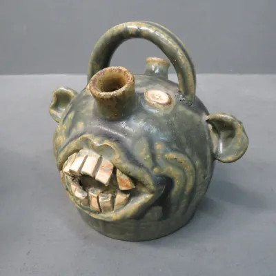 Sale Photo Thumbnail #6: Southern folk art pottery face jug with a green glaze, featuring prominent ceramic teeth, ears, a top handle, and dual spouts.