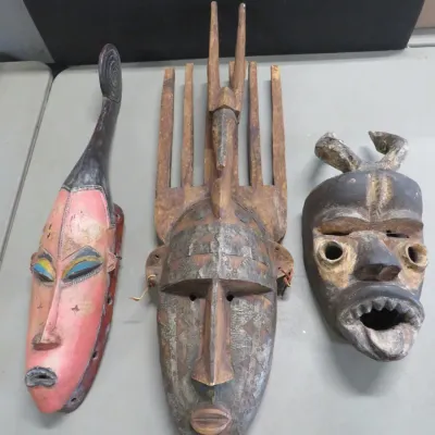 Sale Photo Thumbnail #28: Three hand-carved African tribal masks, including a pink-painted elongated mask, a large mask with a comb-like headdress and metal overlays, and a dark horned mask with an open mouth.