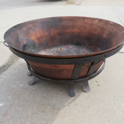 Sale Photo Thumbnail #34: Large copper fire pit bowl resting in a black wrought iron stand, showing signs of previous use and weathering.