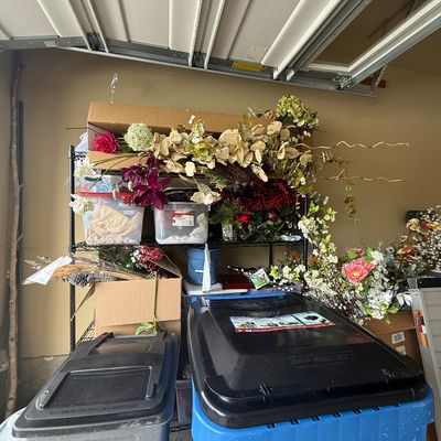 Assorted faux floral stems, greenery, and craft supplies arranged on a black wire shelving unit in a garage.