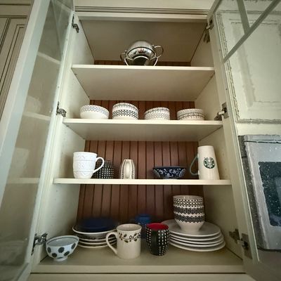 Assorted modern kitchenware, including patterned bowls, mugs, and plates, displayed inside a white cabinet with a wood-paneled back.