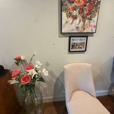 Contemporary upholstered accent chair in a light beige fabric, paired with a large glass floor vase containing faux floral stems and...