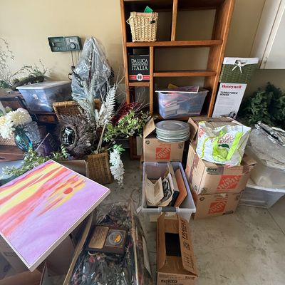 Assorted household items, including a wooden shelving unit, cardboard moving boxes, plastic storage bins, artificial floral arrangements,...