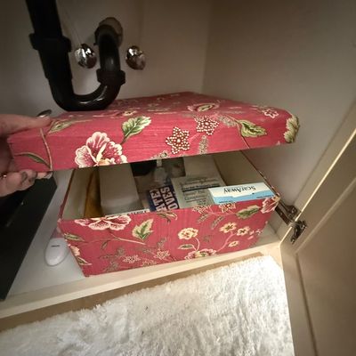 Fabric-covered storage box with a red floral pattern, shown open under a sink with various first aid supplies inside.