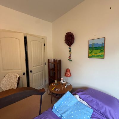 Bedroom corner featuring a round wooden side table with a small lamp, a tall wooden shelf unit, and a desert landscape painting on the wall.