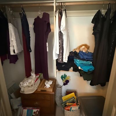 Assorted women's clothing hanging in a closet, including dresses and blouses, alongside a small wooden nightstand, storage baskets,...