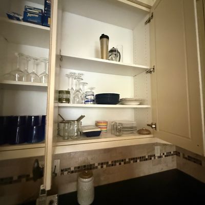 Assorted kitchenware including wine glasses, ceramic plates, bowls, and storage jars, displayed inside cream-colored kitchen cabinets.