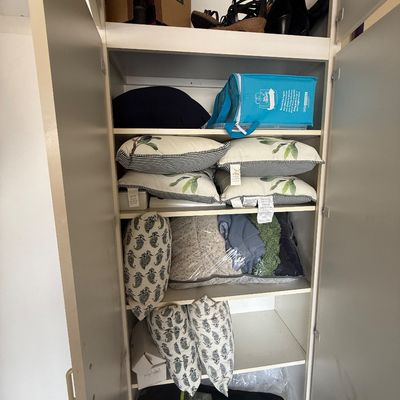 Assorted household linens, throw pillows, and storage items organized on shelves inside a white utility closet.