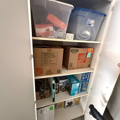 White storage cabinet filled with various items, including plastic storage bins, cardboard moving boxes, and electronics packaging.