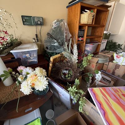 Assorted home decor and storage items, including faux floral arrangements, a wooden bookcase, cardboard boxes, and a colorful abstract...