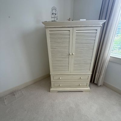 Distressed off-white wooden armoire featuring louvered double doors and two lower drawers with matching wooden knobs.