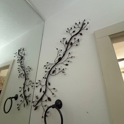 Decorative metal wall art depicting a winding branch with leaves and clear acrylic crystal accents, mounted next to a bathroom mirror.