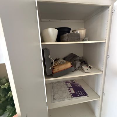 Tall white laminate storage cabinet with adjustable shelves, containing various household items including planters, a vintage metal case,...