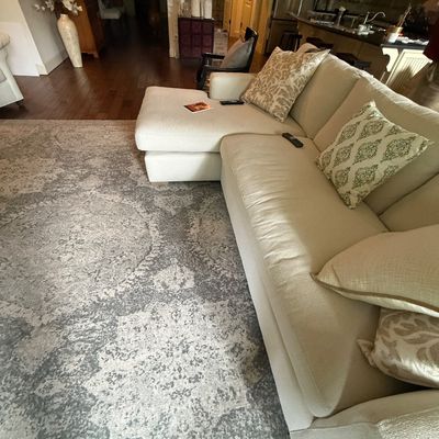 Modern cream-colored sectional sofa with a chaise lounge, accompanied by patterned throw pillows and set on a large grey and white...