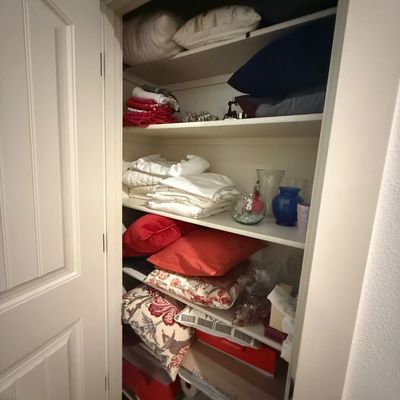 Linen closet filled with assorted bedding, including decorative throw pillows, folded sheets, blankets, and glassware on the shelves.