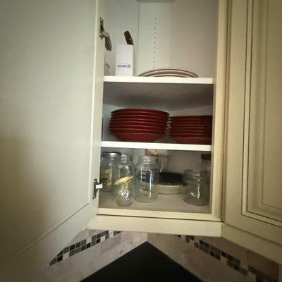 Assorted kitchenware including red ceramic plates, clear glass jars, and white plates stored inside a corner kitchen cabinet.