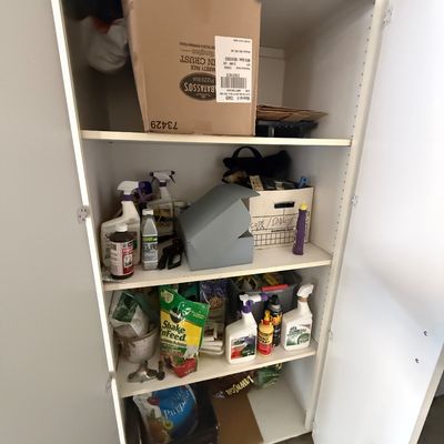White laminate storage cabinet filled with various household items, including lawn care products, cleaning sprays, and cardboard boxes.