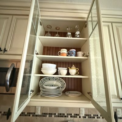 Assorted kitchenware including plates, bowls, mugs, and glassware, displayed inside an open cream-colored cabinet with glass doors.