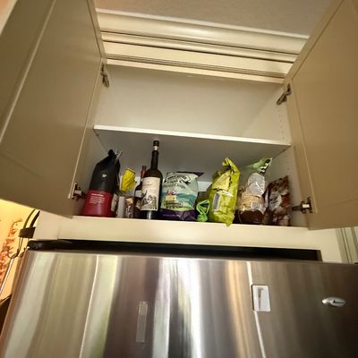 Amana stainless steel refrigerator positioned beneath a beige double-door kitchen cabinet containing various pantry items and bottles.