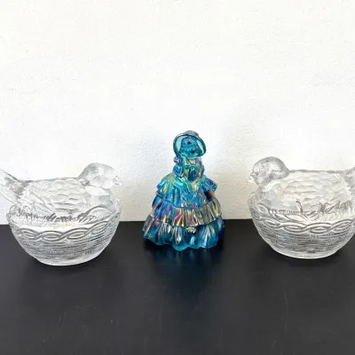 Sale Photo Thumbnail #338: Fenton blue iridescent carnival glass Southern Belle figurine flanked by a pair of vintage clear glass bird-on-nest covered dishes.