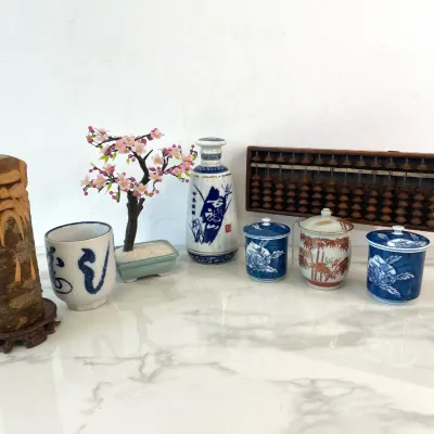 Sale Photo Thumbnail #70: Collection of Asian decorative items including a vintage wooden abacus, a hand-carved wood spirit sculpture on a stand, a faux cherry blossom bonsai tree, and various blue and white ceramic tea vessels and sake bottles.