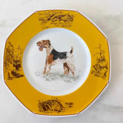 Sale Photo Thumbnail #177: Hermes porcelain plate from the 'Chasse a Courre' collection, featuring a Wire Hair Fox Terrier illustration in the center and a yellow rim decorated with hunting scenes.