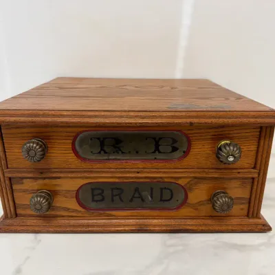 Sale Photo Thumbnail #228: Antique oak spool cabinet featuring two drawers with glass inserts reading 'R. B.' and 'BRAID', complete with original metal pull knobs.