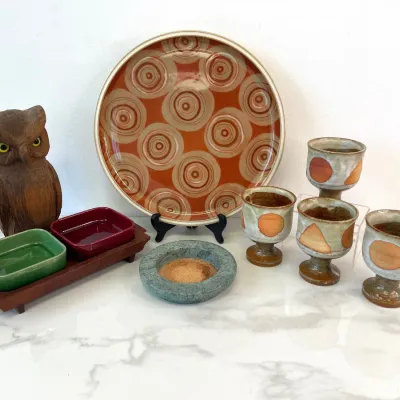 Sale Photo Thumbnail #69: Mid-century modern stoneware collection featuring a circular platter with geometric concentric circles, four matching footed goblets, a small green stone dish, a wooden owl figurine, and a teak tray with red and green ceramic inserts.