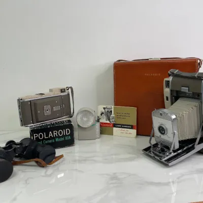 Sale Photo Thumbnail #420: Vintage Polaroid Land Camera Model 80A with its original box, a Polaroid Model 150 folding bellows instant camera with a brown leather carrying case, a flash attachment, manual, and a pair of vintage black binoculars.