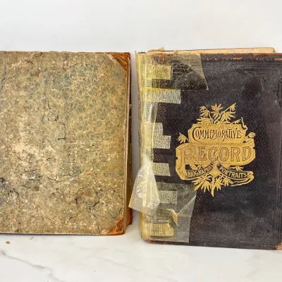 Sale Photo Thumbnail #208: Two antique 19th-century books, including a heavily worn 'Commemorative Record of Biographies and Portraits' with a gold-embossed cover, showing significant wear, tape repairs, and aged patina.