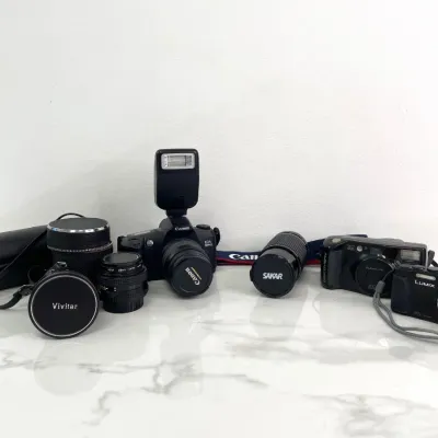 Sale Photo Thumbnail #393: Vintage camera gear collection including a Canon EOS Rebel SLR camera with flash, a Panasonic Lumix digital camera, a Fuji Discovery point-and-shoot, and various lenses by Vivitar, Sakar, and Canon.