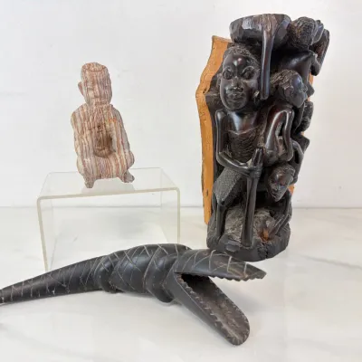 Sale Photo Thumbnail #101: Collection of hand-carved tribal art, featuring a Makonde-style African family tree sculpture in dark wood, a carved wooden crocodile, and a small stone figure.