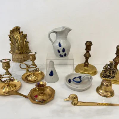 Sale Photo Thumbnail #86: Collection of vintage brass and stoneware decor, featuring brass chambersticks, candlesticks, a brass duck letter opener, and a brass bell alongside blue-and-grey salt-glazed stoneware pottery pieces.