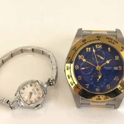 Sale Photo Thumbnail #302: Vintage Gruen Precision ladies' cocktail watch with a silver-tone stretch band alongside a Stauer 27 Jewels automatic chronograph watch featuring a blue dial and gold-tone bezel.