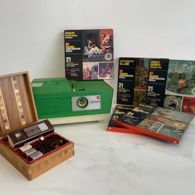 Sale Photo Thumbnail #154: Vintage GAF Talking View-Master projector in green and white, accompanied by several GAF Talking View-Master reel sets including Rudolph, G.I. Joe, and Charlie Brown, alongside a vintage Tru-Vue stereoscope viewer in a wooden storage box.