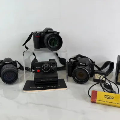 Sale Photo Thumbnail #175: Vintage camera collection featuring three Nikon DSLR cameras with lenses, a Polaroid Pronto! Land Camera with manual, a Cine-Kodak Eight-25 movie camera, and a Milo lens cleaning brush.