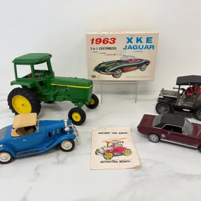 Sale Photo Thumbnail #84: Vintage toy collection featuring a green John Deere tractor, a blue Hubley roadster, a maroon Ford Mustang, a classic Model T, and a 1963 Monogram XKE Jaguar model kit box.